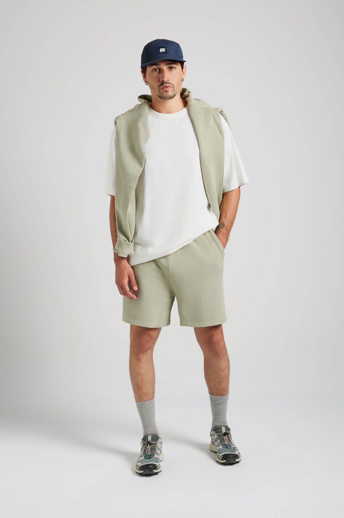 Persona che indossa gli Essential Sunwashed Sweatshorts in Seagrass, abbinati a una camicia bianca e sneakers casual, in piedi con le mani in tasca | TP_IMAGE_TYPE=MODEL_FRONT