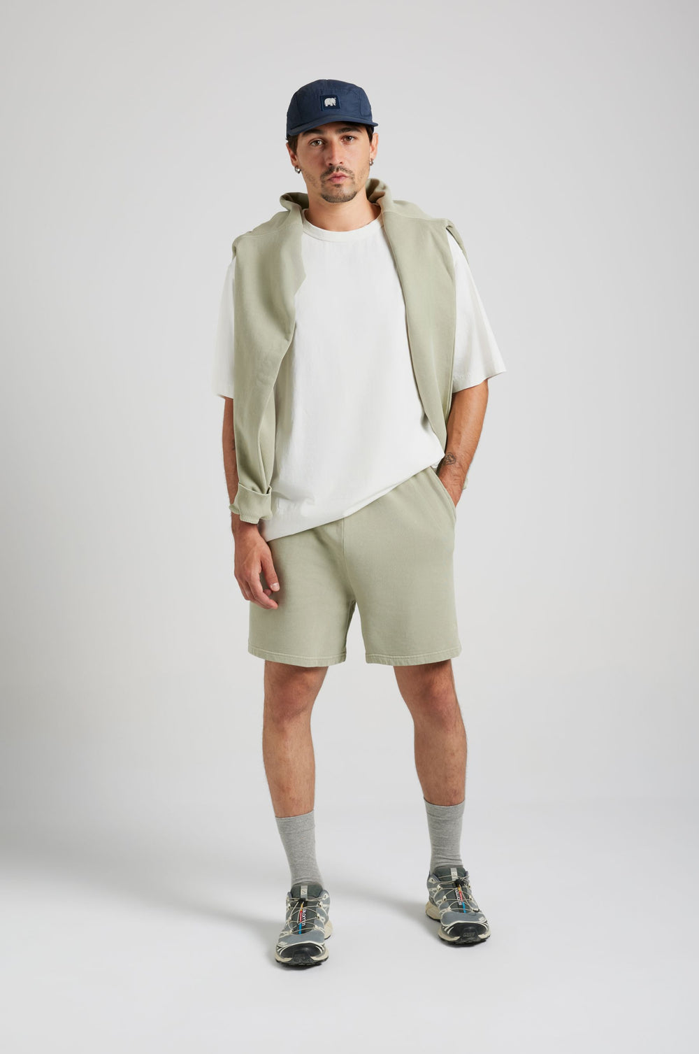 Persona che indossa gli Essential Sunwashed Sweatshorts in Seagrass, abbinati a una camicia bianca e sneakers casual, in piedi con le mani in tasca | TP_IMAGE_TYPE=MODEL_FRONT