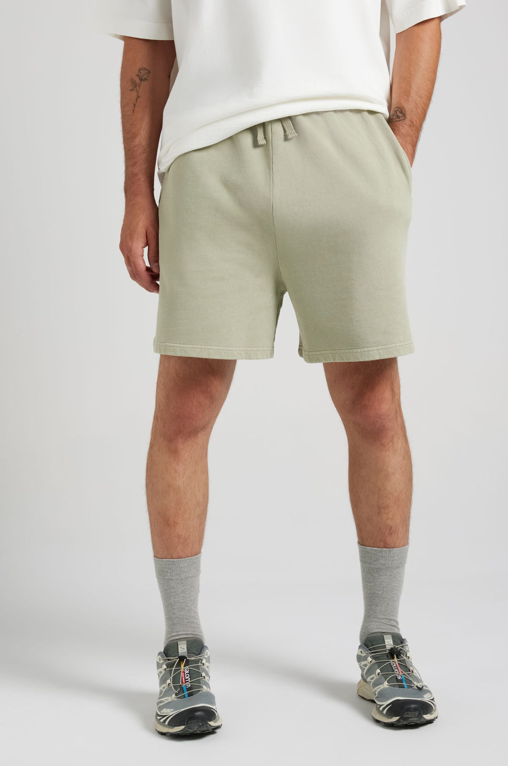 Persona che indossa Essential Sunwashed Sweatshorts in Seagrass, abbinati a calzini grigi e sneakers, in piedi su uno sfondo neutro | TP_IMAGE_TYPE=MODEL_FRONT