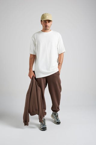 Persona che indossa gli Essential Sweatpants in Shaved Chocolate, abbinati a una t-shirt bianca e a un cappellino beige, in piedi davanti a uno sfondo neutro. | TP_IMAGE_TYPE=MODEL_FRONT