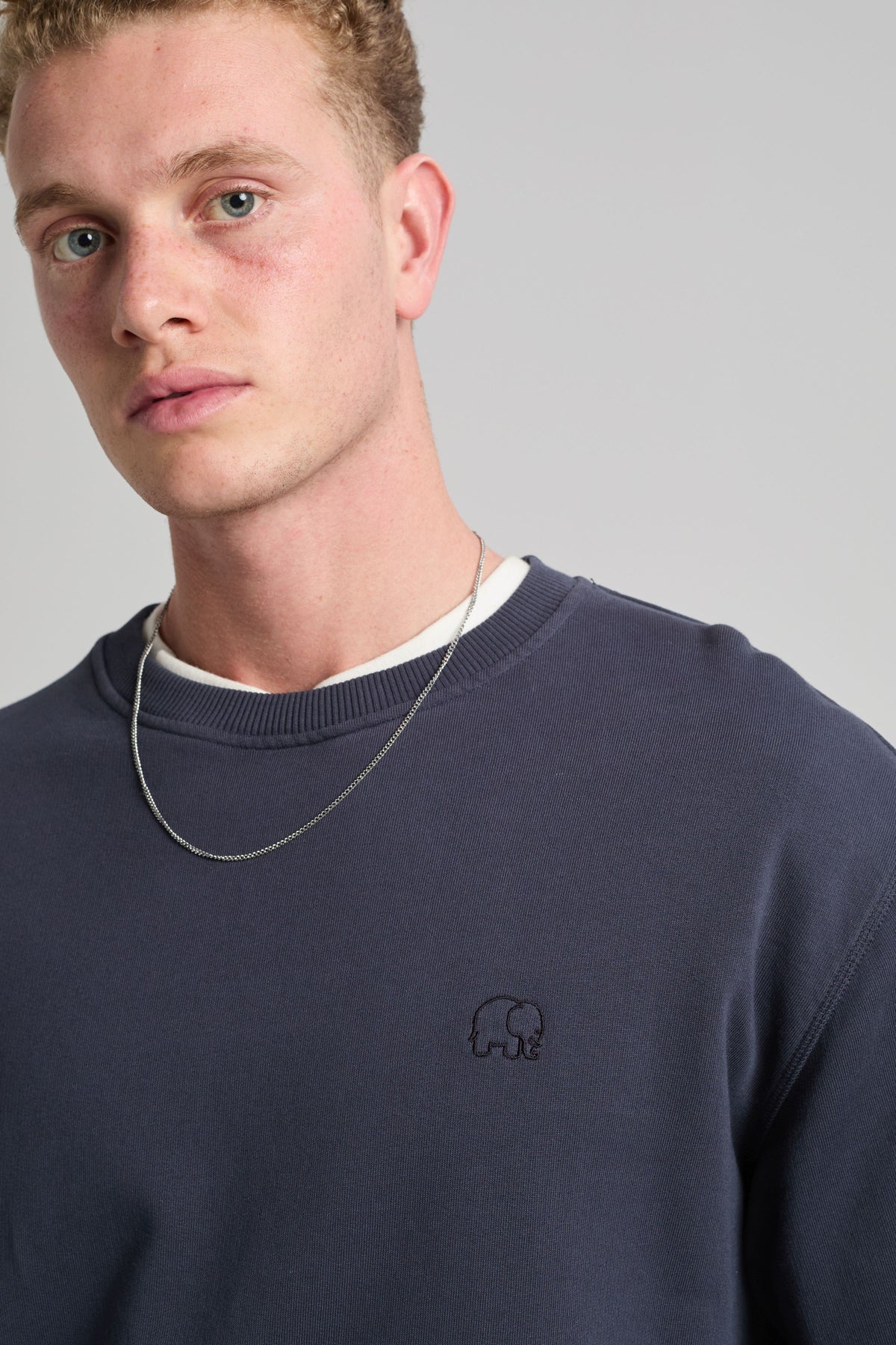 Persona che indossa una Essential Sweatshirt in Parisian Blue, con un logo elefante ricamato, abbinata a una collana a catena argentata. | TP_IMAGE_TYPE=MODEL_FRONT
