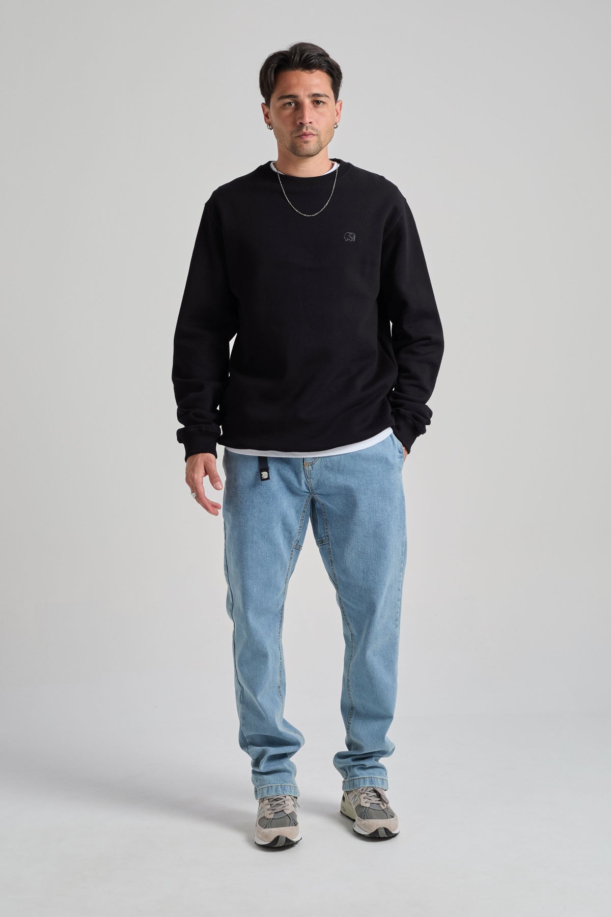 Persona che indossa una Organic Essential Sweatshirt nera, abbinata a jeans azzurri e sneakers, in piedi davanti a uno sfondo neutro. | TP_IMAGE_TYPE=MODEL_FRONT
