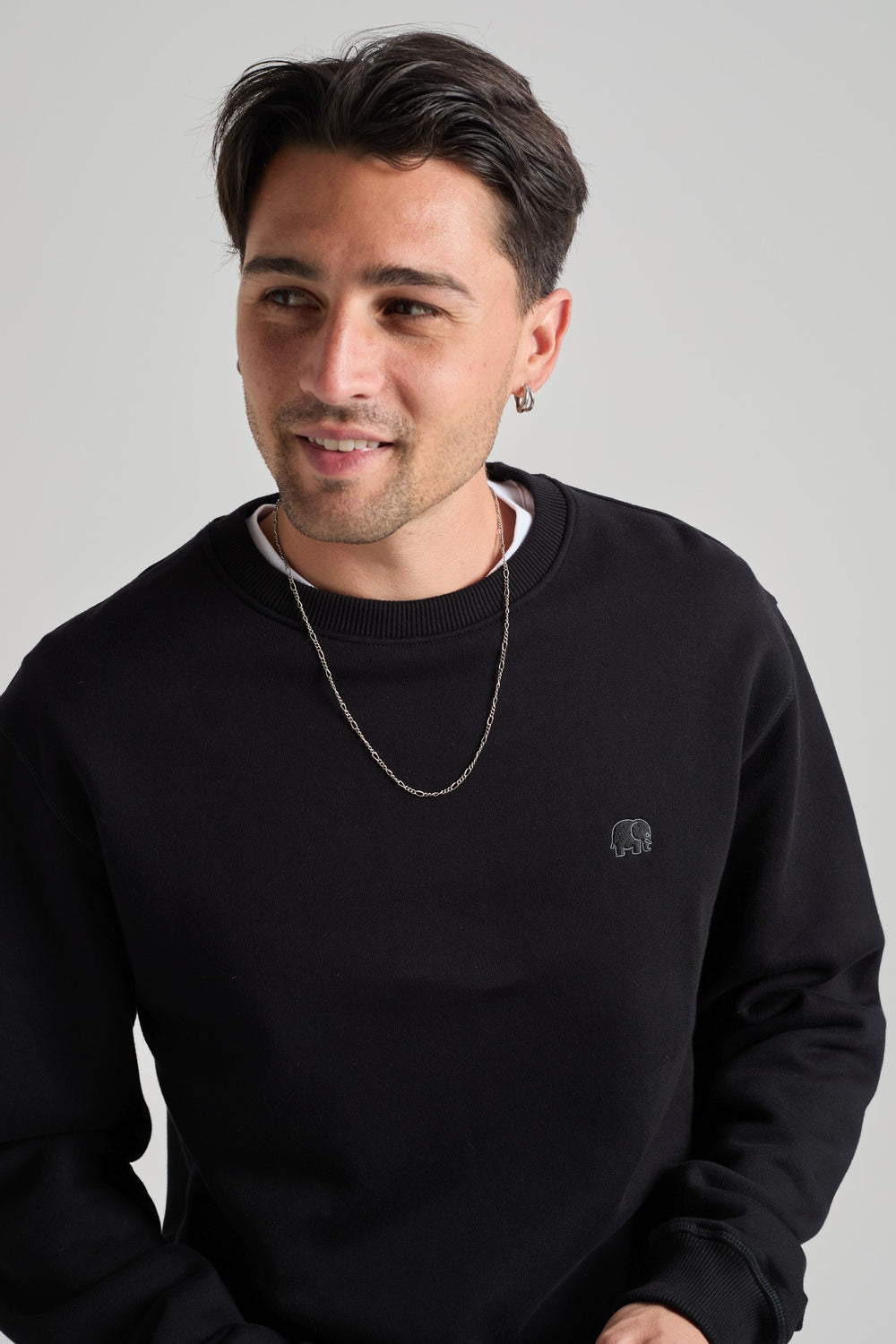 Persona che indossa una Organic Essential Sweatshirt Black con un piccolo logo a forma di elefante, abbinata a una collana con catena argentata, mentre sorride su uno sfondo neutro. | TP_IMAGE_TYPE=MODEL_FRONT