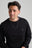 Persona che indossa una Organic Essential Sweatshirt Black con un piccolo logo a forma di elefante, abbinata a una collana con catena argentata, mentre sorride su uno sfondo neutro. | TP_IMAGE_TYPE=MODEL_FRONT