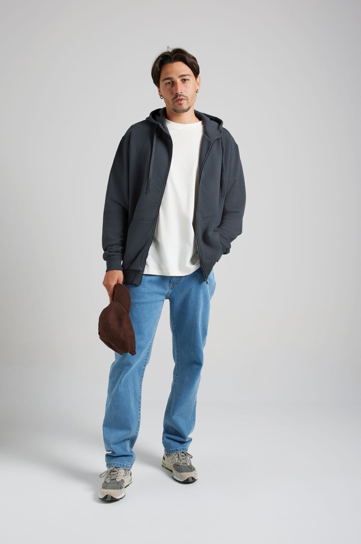 Personne portant un sweat à capuche Essential Zip en Bitumen Grey, associé à une chemise blanche et un jean bleu, tenant un chapeau marron | TP_IMAGE_TYPE=MODEL_FRONT