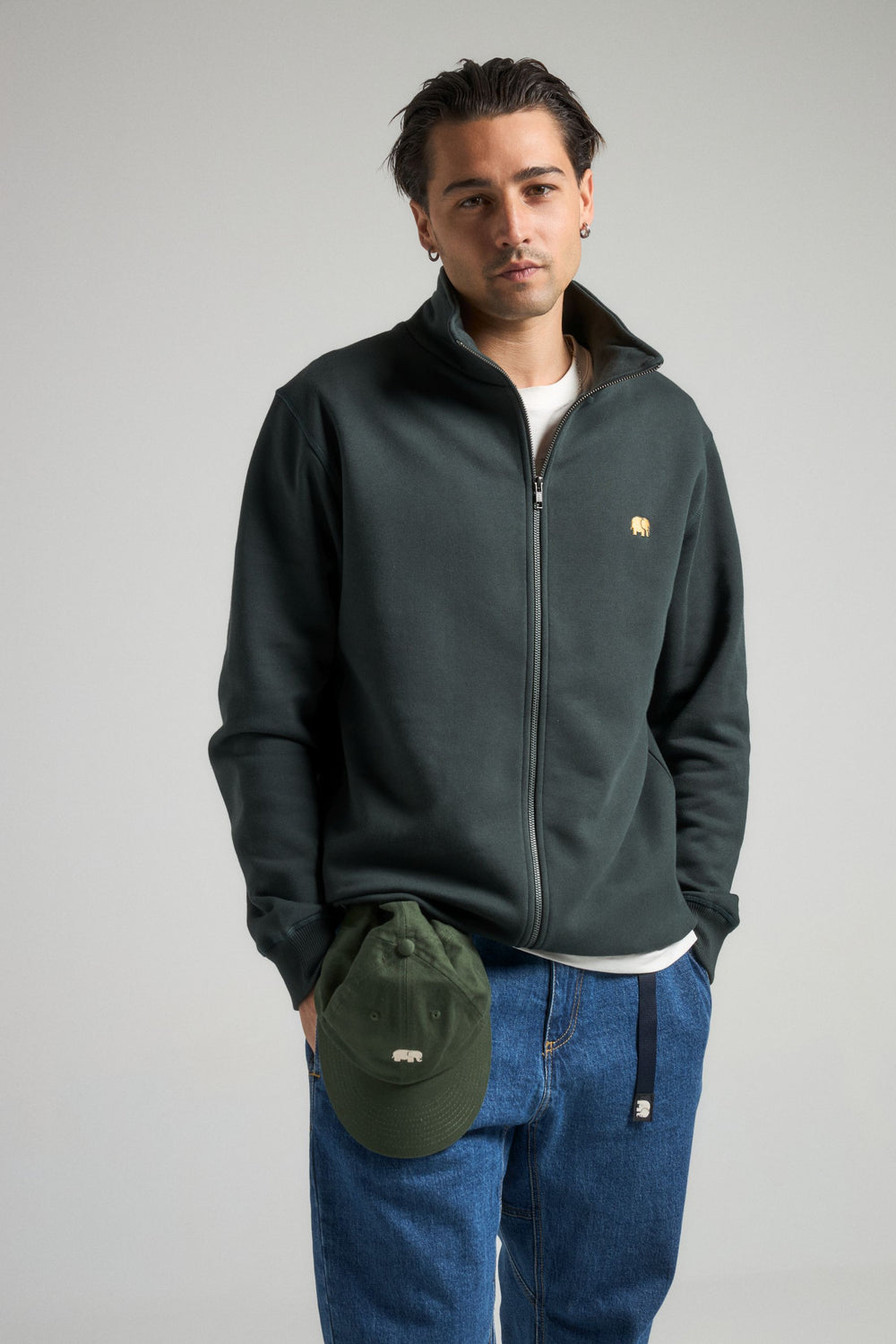 Personne portant un Organic Essential Zip Sweatshirt en Scarab Green, associé à une casquette verte assortie et à un jean bleu, debout devant un fond neutre. | TP_IMAGE_TYPE=MODEL_FRONT