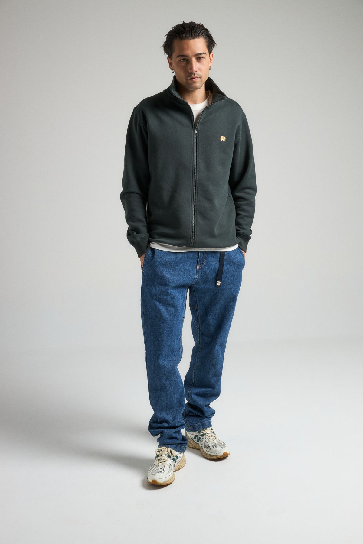 Personne portant un Organic Essential Zip Sweatshirt en Scarab Green, associé à un jean décontracté et des baskets, se tenant avec assurance sur un fond neutre. | TP_IMAGE_TYPE=MODEL_FRONT