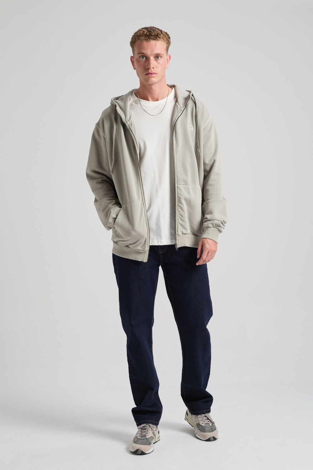 Persona che indossa una Essential Sunwashed Zip Hoodie in Natural Gray, abbinata a una t-shirt bianca e a jeans scuri, in posa su uno sfondo uniforme. | TP_IMAGE_TYPE=MODEL_FRONT