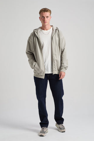 Persona che indossa una Essential Sunwashed Zip Hoodie in Natural Gray, abbinata a una t-shirt bianca e a jeans scuri, in posa su uno sfondo uniforme. | TP_IMAGE_TYPE=MODEL_FRONT