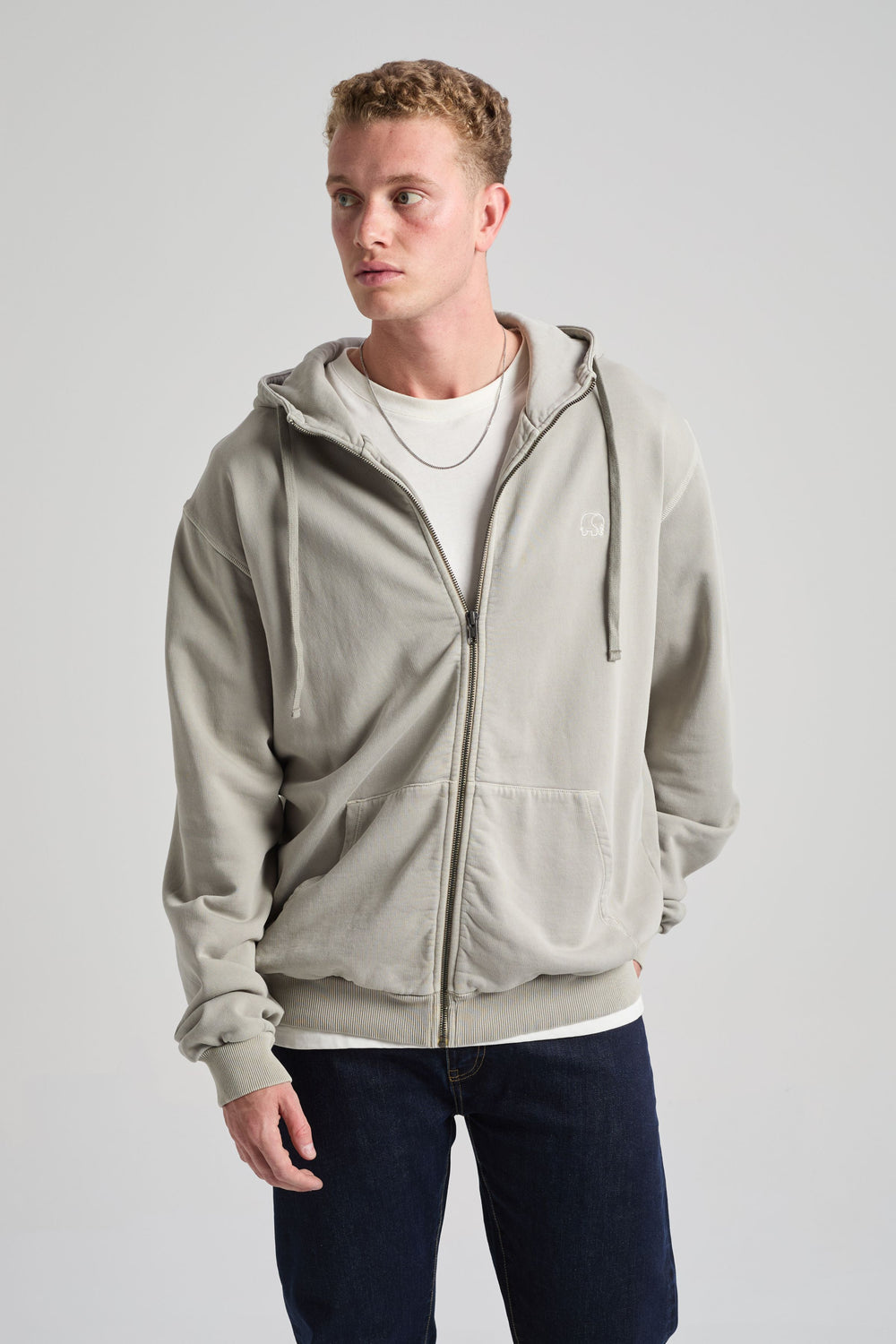 Persona che indossa la Essential Sunwashed Zip Hoodie in Natural Gray, abbinata a una t-shirt bianca e a jeans scuri, in posa in uno studio fotografico. | TP_IMAGE_TYPE=MODEL_FRONT