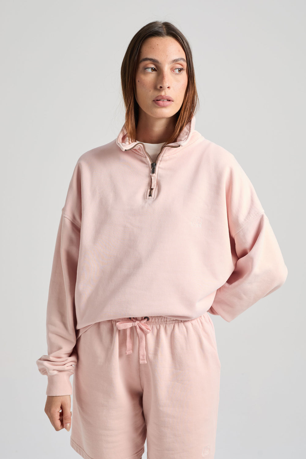 Persona che indossa una Sunwashed Essential Quarter Zip Sweatshirt da donna in lotus pink, abbinata a shorts coordinati, con lo sguardo rivolto di lato su sfondo neutro. | TP_IMAGE_TYPE=MODEL_FRONT