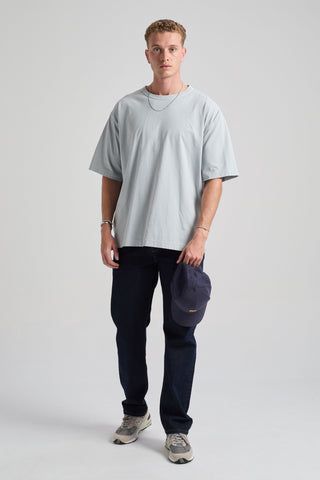 Personne portant un t-shirt oversize bleu perle à manches courtes, associé à un jean en denim foncé et tenant une casquette bleu marine, dans un studio. | TP_IMAGE_TYPE=MODEL_FRONT