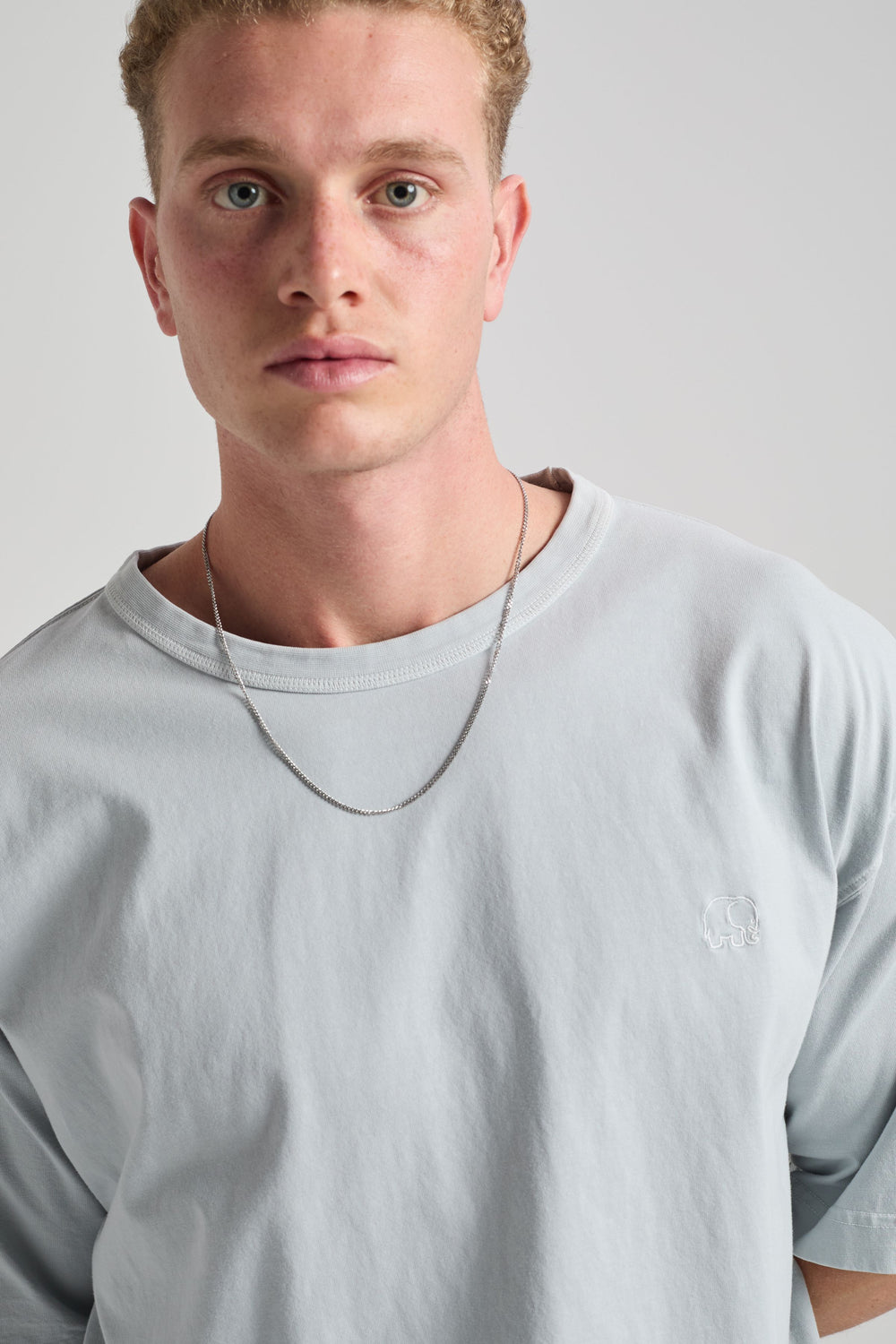Personne portant un Essential Sunwashed Heavyweight Oversized T-Shirt dans le coloris Pearl Blue, avec un look décontracté et un collier à chaîne argentée. | TP_IMAGE_TYPE=MODEL_FRONT