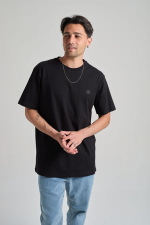Personne portant un T-Shirt Organic Essential noir, associé à un jean bleu clair et une chaîne argentée, dans une pose décontractée. | TP_IMAGE_TYPE=MODEL_FRONT