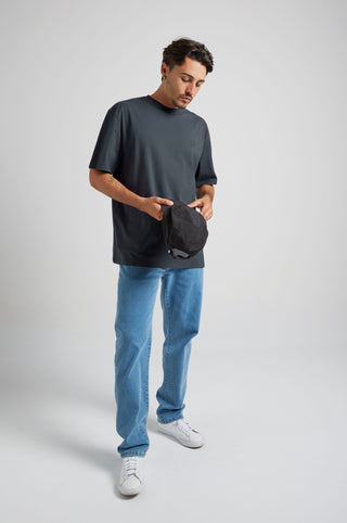 Person wearing an Essential T-Shirt in Bitumen Grey, holding a hat while standing in a casual pose. | TP_IMAGE_TYPE=MODEL_FRONT