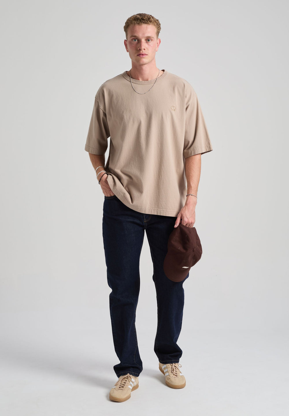 Personne portant un Essential Oversized T-Shirt en Moon Rock Grey, avec un jean foncé et tenant une casquette marron | TP_IMAGE_TYPE=MODEL_FRONT