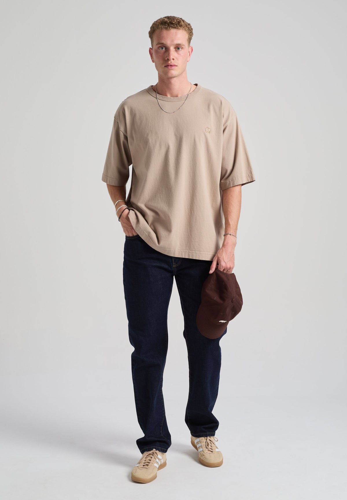 Personne portant un Essential Oversized T-Shirt en Moon Rock Grey, avec un jean foncé et tenant une casquette marron | TP_IMAGE_TYPE=MODEL_FRONT