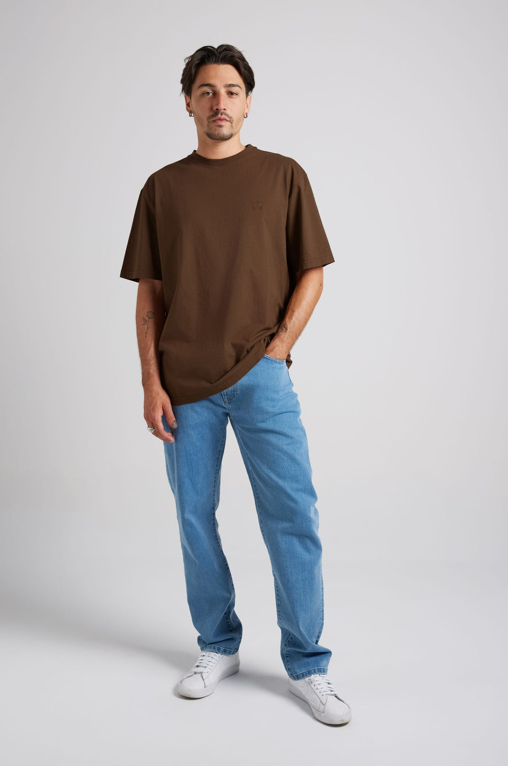 Person wearing an Essential T-Shirt in Carafe Brown paired with light blue jeans and white sneakers, standing in a relaxed pose. | TP_IMAGE_TYPE=MODEL_FRONT