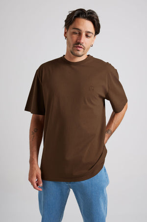Person wearing Essential T-Shirt in Carafe Brown, styled with light blue jeans, posing in a neutral background. | TP_IMAGE_TYPE=MODEL_FRONT