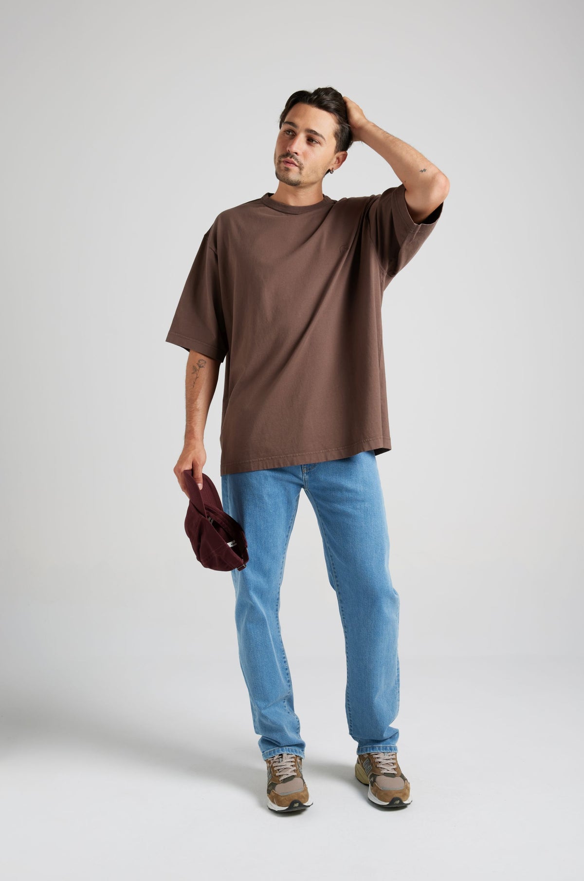 Personne portant un t-shirt oversize dans un riche coloris chocolat, associé à un jean bleu clair et tenant une casquette bordeaux. | TP_IMAGE_TYPE=MODEL_FRONT
