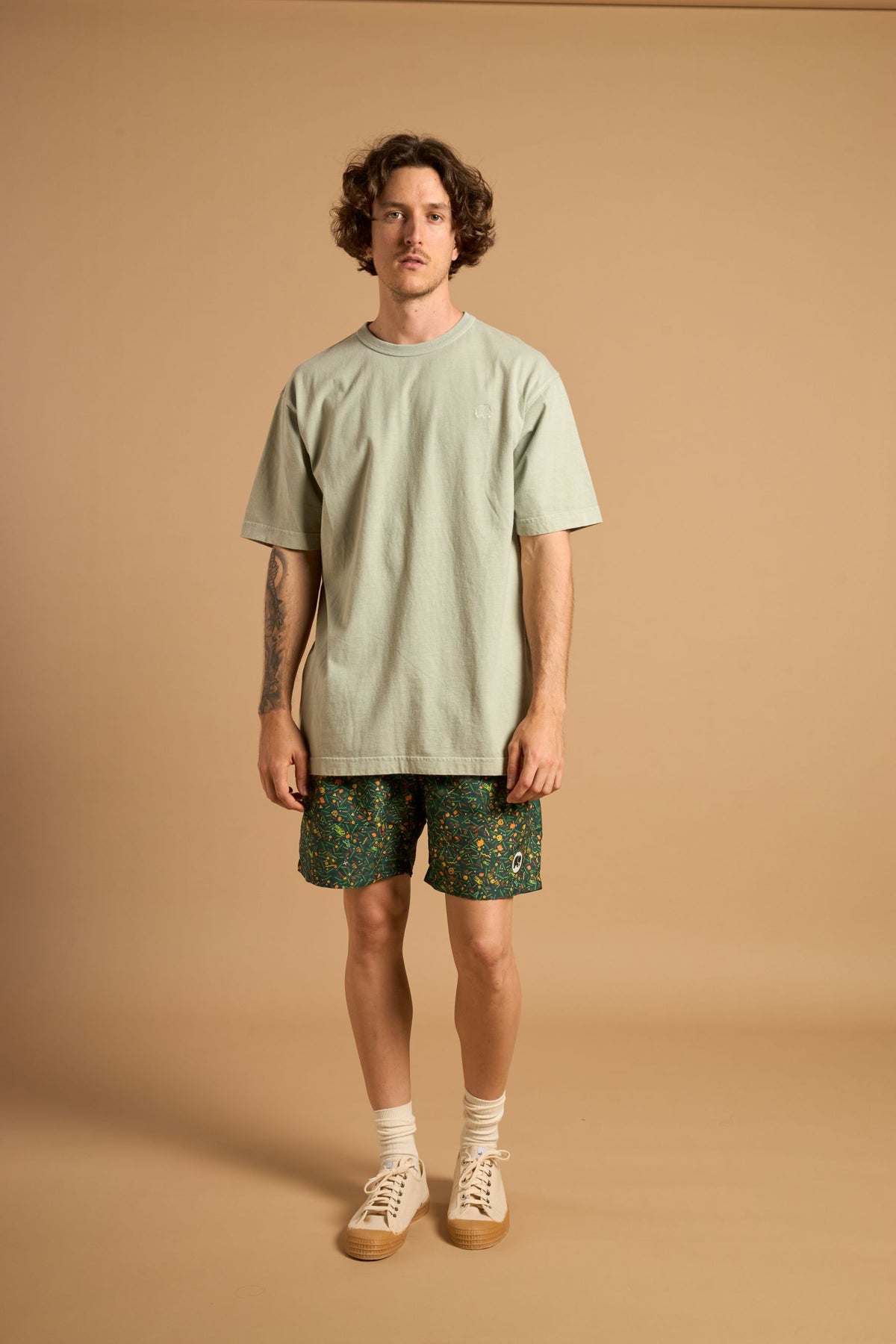 Personne portant un Dogtown SEAQUAL®YARN Swim Trunks vert, associé à un t-shirt vert clair et à des baskets beiges, debout sur un fond beige. | TP_IMAGE_TYPE=MODEL_FRONT
