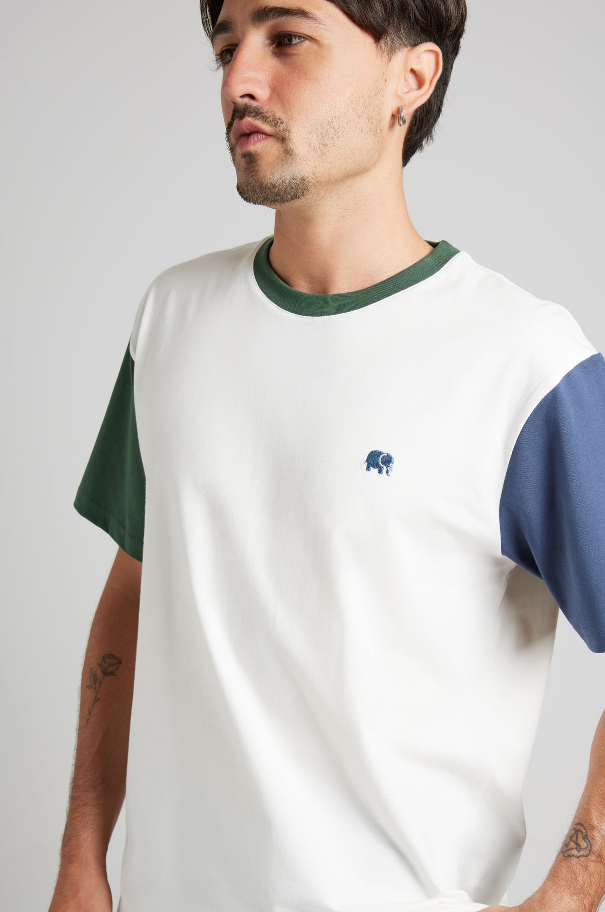 Persona che indossa una t-shirt Color Block in cotone organico con tonalità bianco naturale, verde e blu, che mostra un delicato logo elefante sul petto. | TP_IMAGE_TYPE=MODEL_FRONT