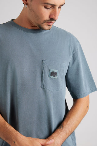 Personne portant un t-shirt Garza Pigment Dyed en Elm Green avec un détail de poche et un design de logo, le regard légèrement baissé. | TP_IMAGE_TYPE=MODEL_FRONT