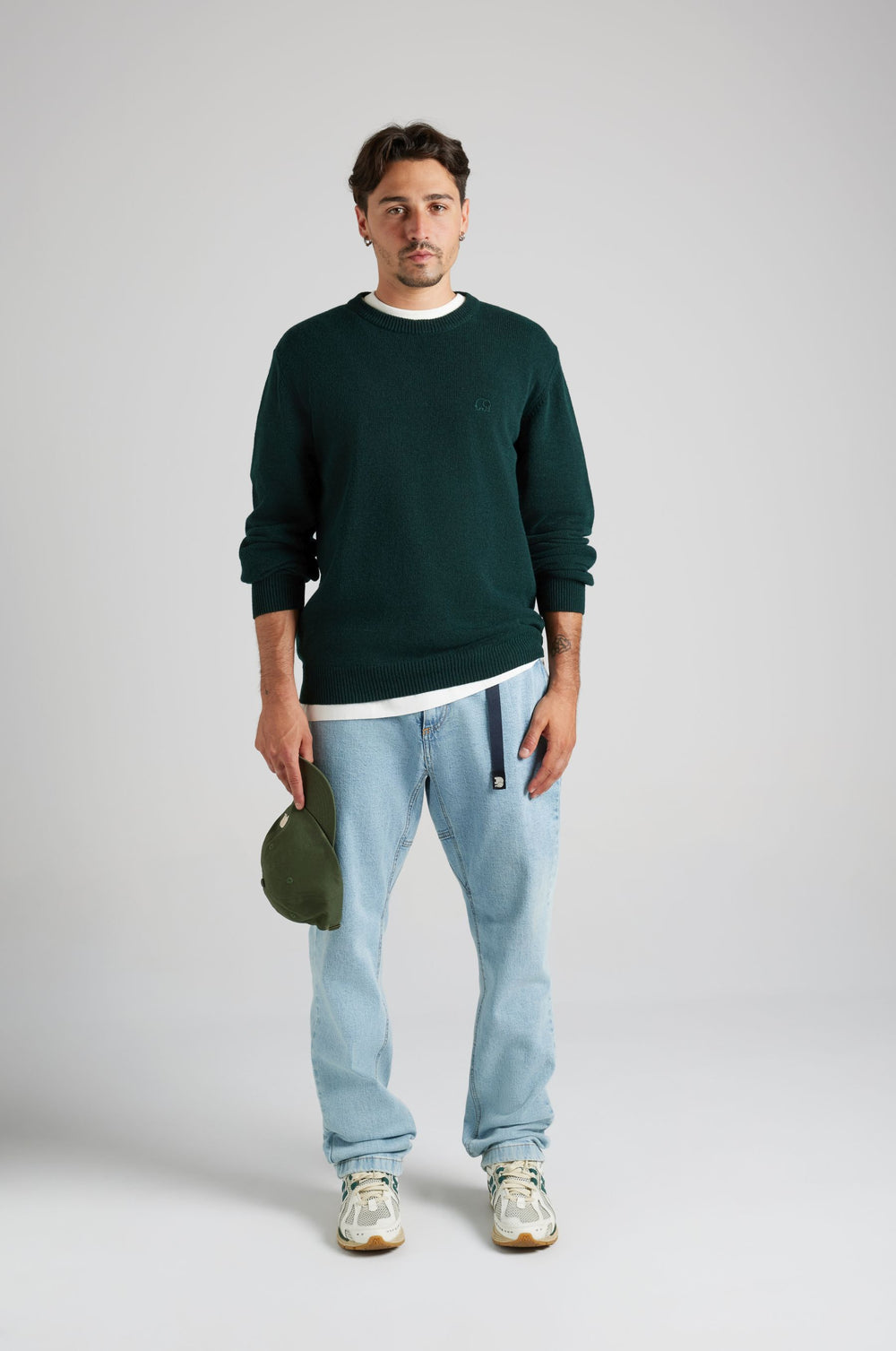 Persona che indossa il Tarbena Knit in Pine Green, abbinato a una maglietta bianca e a jeans azzurri, con un cappellino verde in mano. | TP_IMAGE_TYPE=MODEL_FRONT