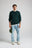 Persona che indossa il Tarbena Knit in Pine Green, abbinato a una maglietta bianca e a jeans azzurri, con un cappellino verde in mano. | TP_IMAGE_TYPE=MODEL_FRONT