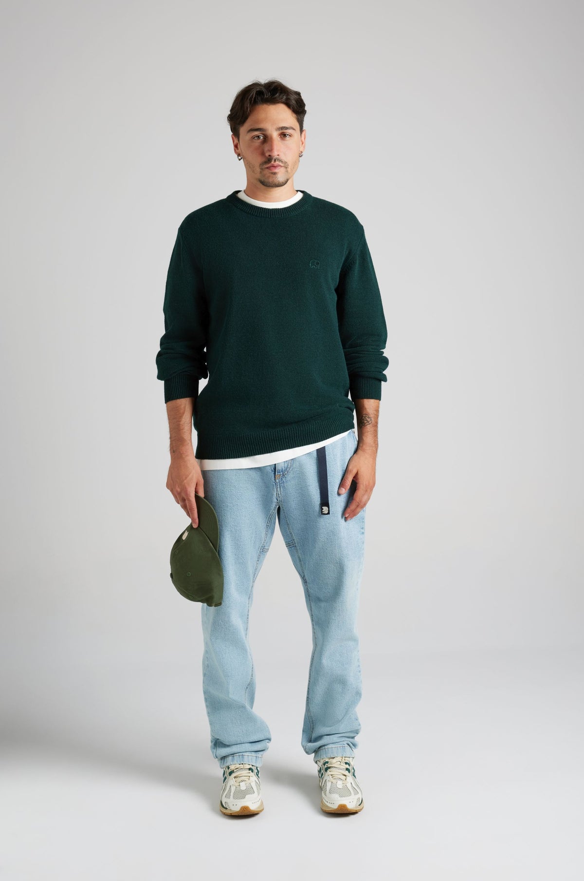 Persona che indossa il Tarbena Knit in Pine Green, abbinato a una maglietta bianca e a jeans azzurri, con un cappellino verde in mano. | TP_IMAGE_TYPE=MODEL_FRONT