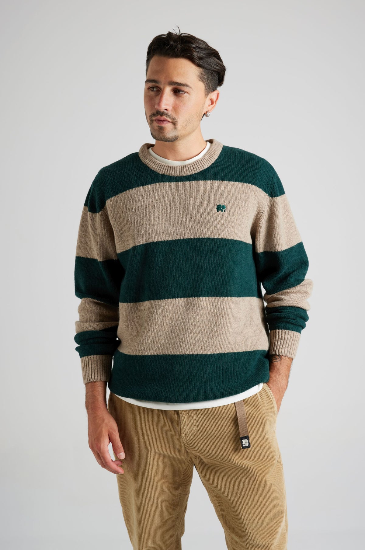 Persona che indossa un maglione Sella Knit in pine green e nomad brown, con righe e logo ricamato, abbinato a pantaloni in velluto a coste beige. | TP_IMAGE_TYPE=MODEL_FRONT