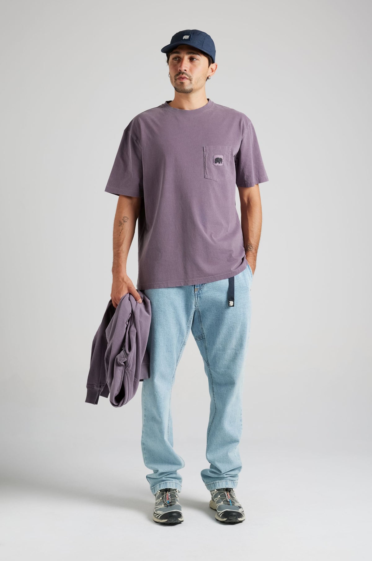 Persona che indossa la Garza Pigment Dyed T-Shirt in Sunset Purple, con in mano un capo abbinato, abbinata a pantaloni azzurri e un cappello | TP_IMAGE_TYPE=MODEL_FRONT