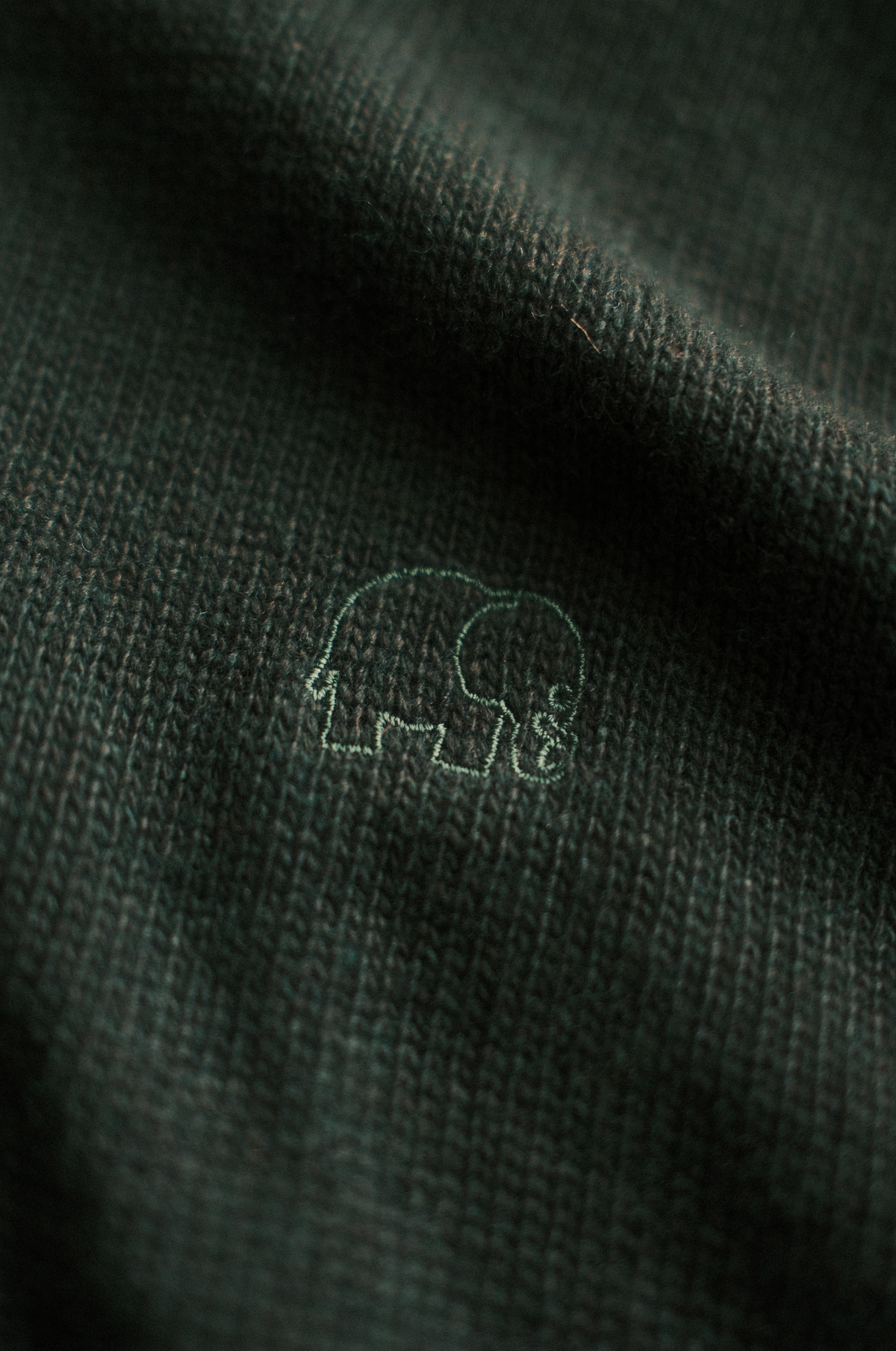 Dettaglio ravvicinato del Tarbena Knit in Pine Green che mostra lo scollo a coste e il logo dell'elefante. | TP_IMAGE_TYPE=DETAIL