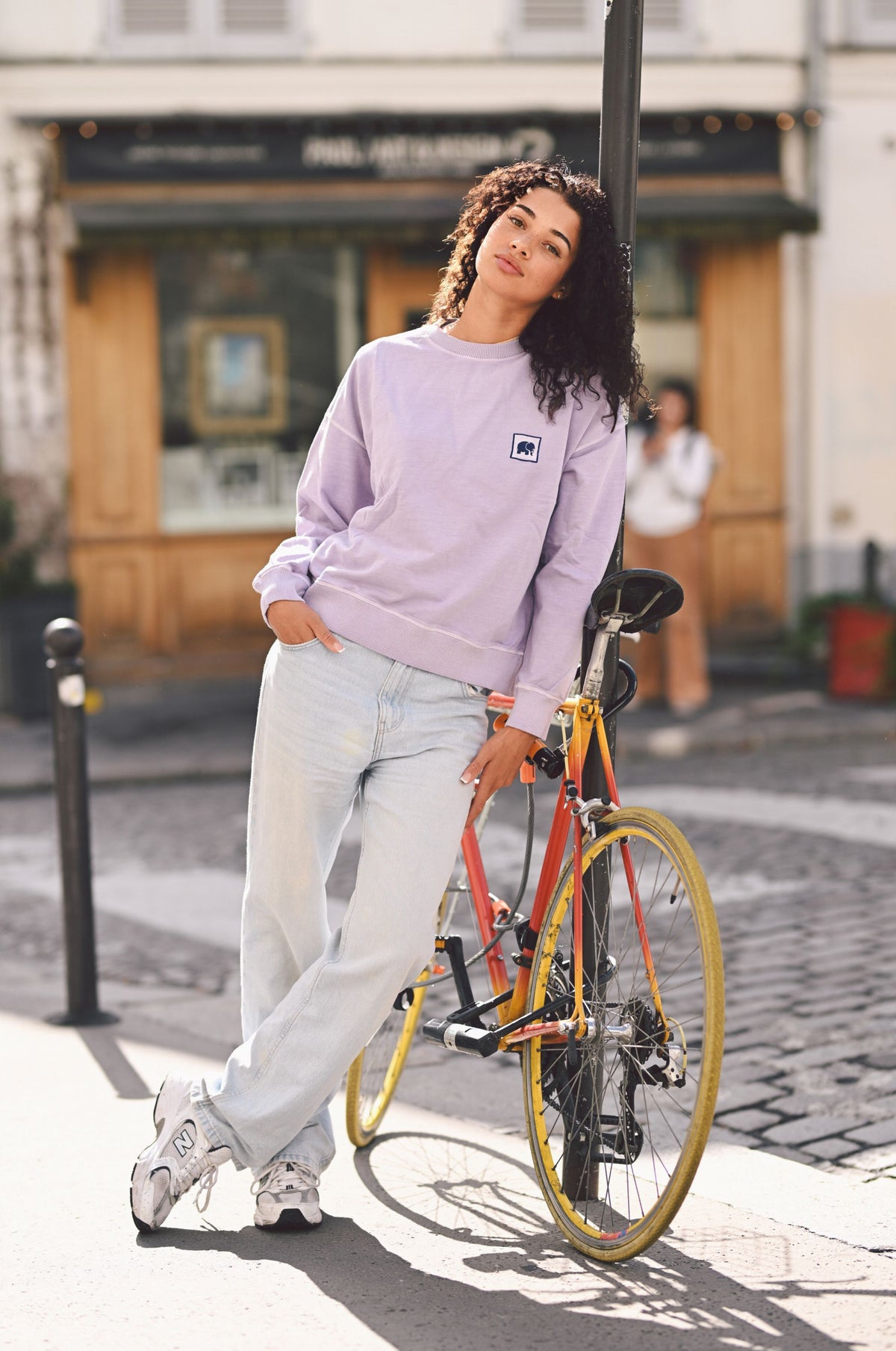 Personne portant un sweat-shirt oversize lavande dans un cadre extérieur, appuyée contre un vélo | TP_IMAGE_TYPE=MODEL_FRONT