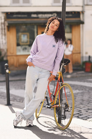 Personne portant un sweat-shirt oversize lavande dans un cadre extérieur, appuyée contre un vélo | TP_IMAGE_TYPE=MODEL_FRONT