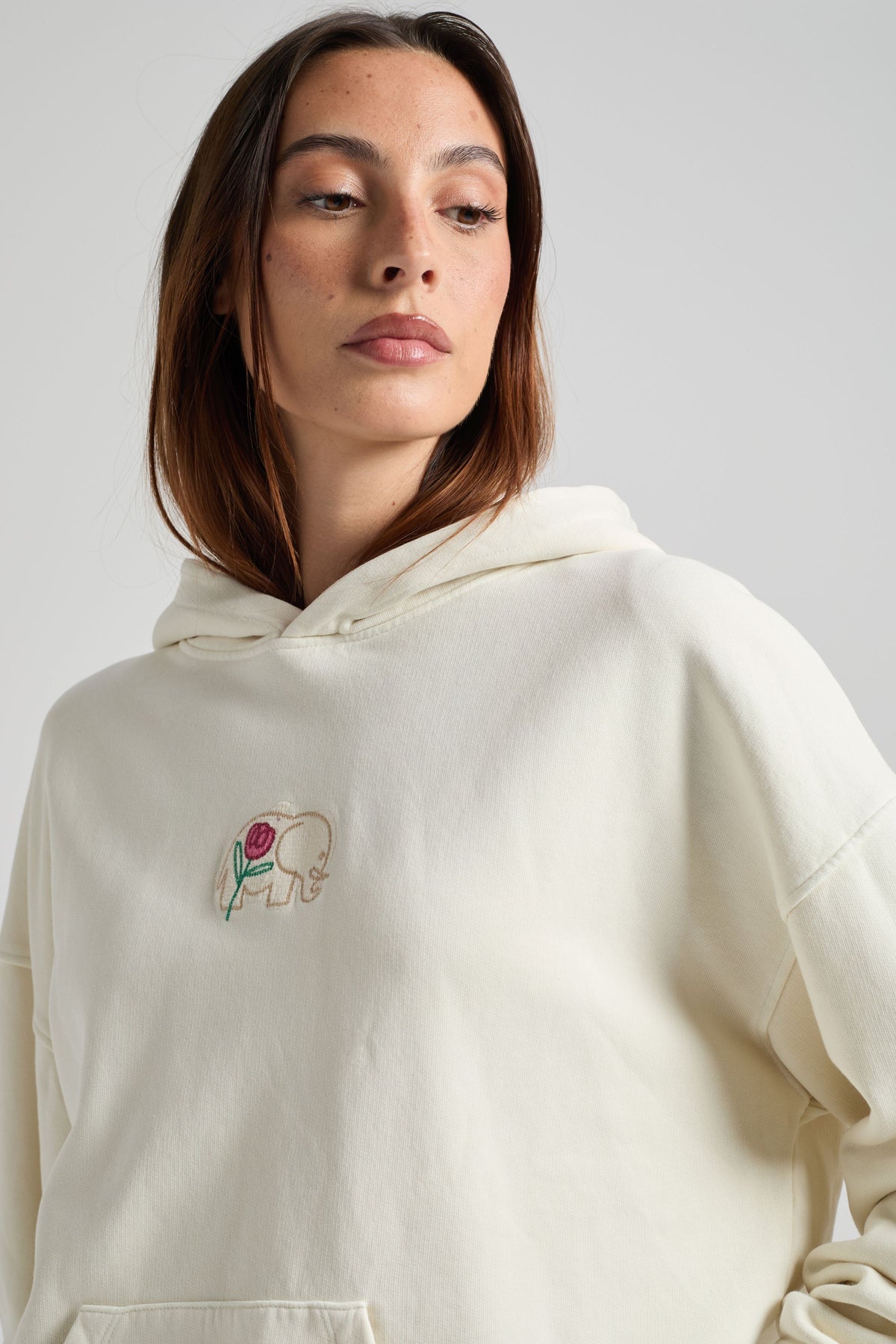 Personne portant un sweat à capuche femme blanc antique avec une broderie d'éléphant sur la poitrine, regardant sur le côté. | TP_IMAGE_TYPE=MODEL_FRONT
