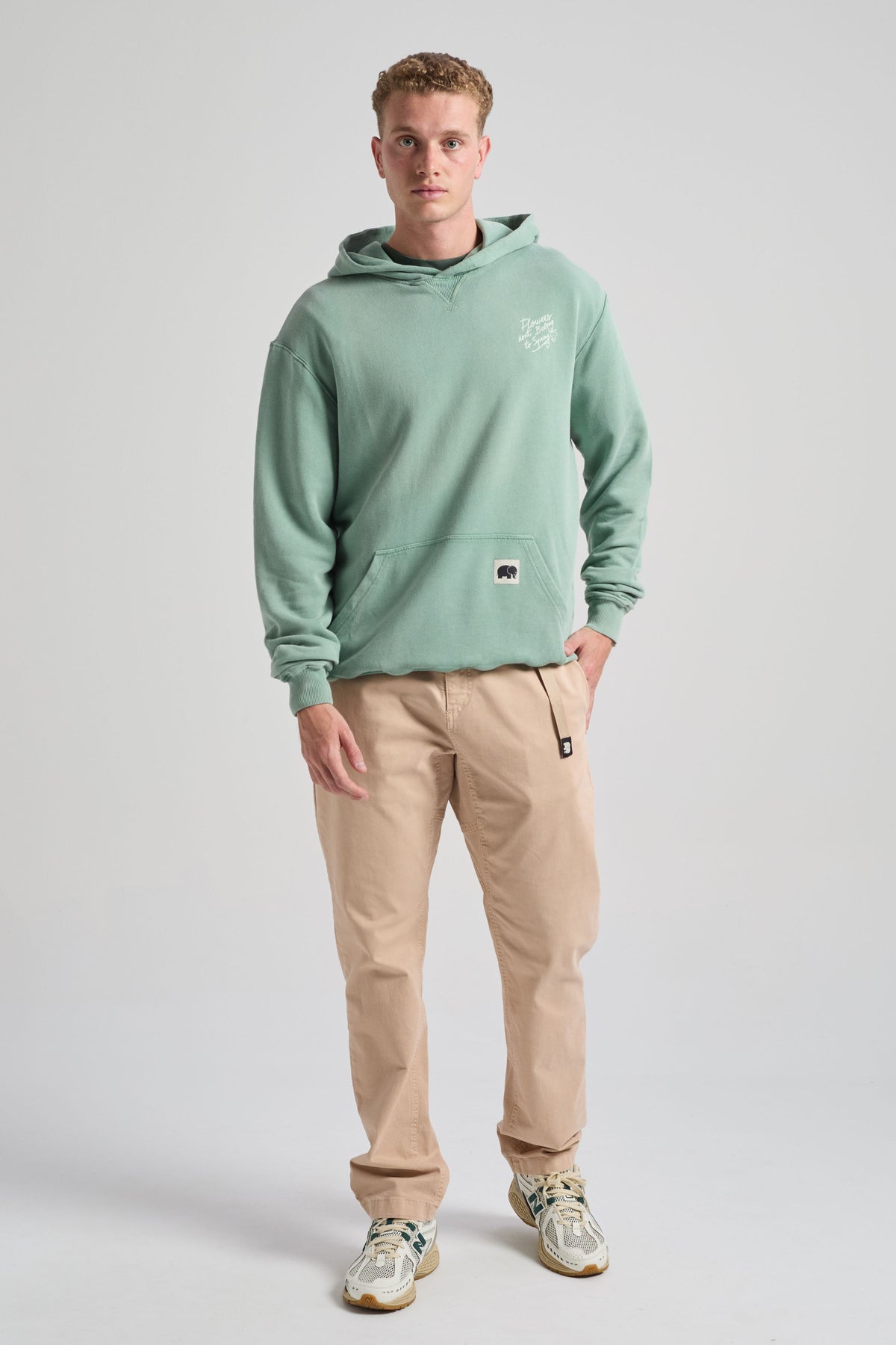 Persona che indossa una Flowers Hoodie in Vintage Sage con tasca frontale, abbinata a pantaloni beige e sneakers, in piedi davanti a uno sfondo neutro | TP_IMAGE_TYPE=MODEL_FRONT