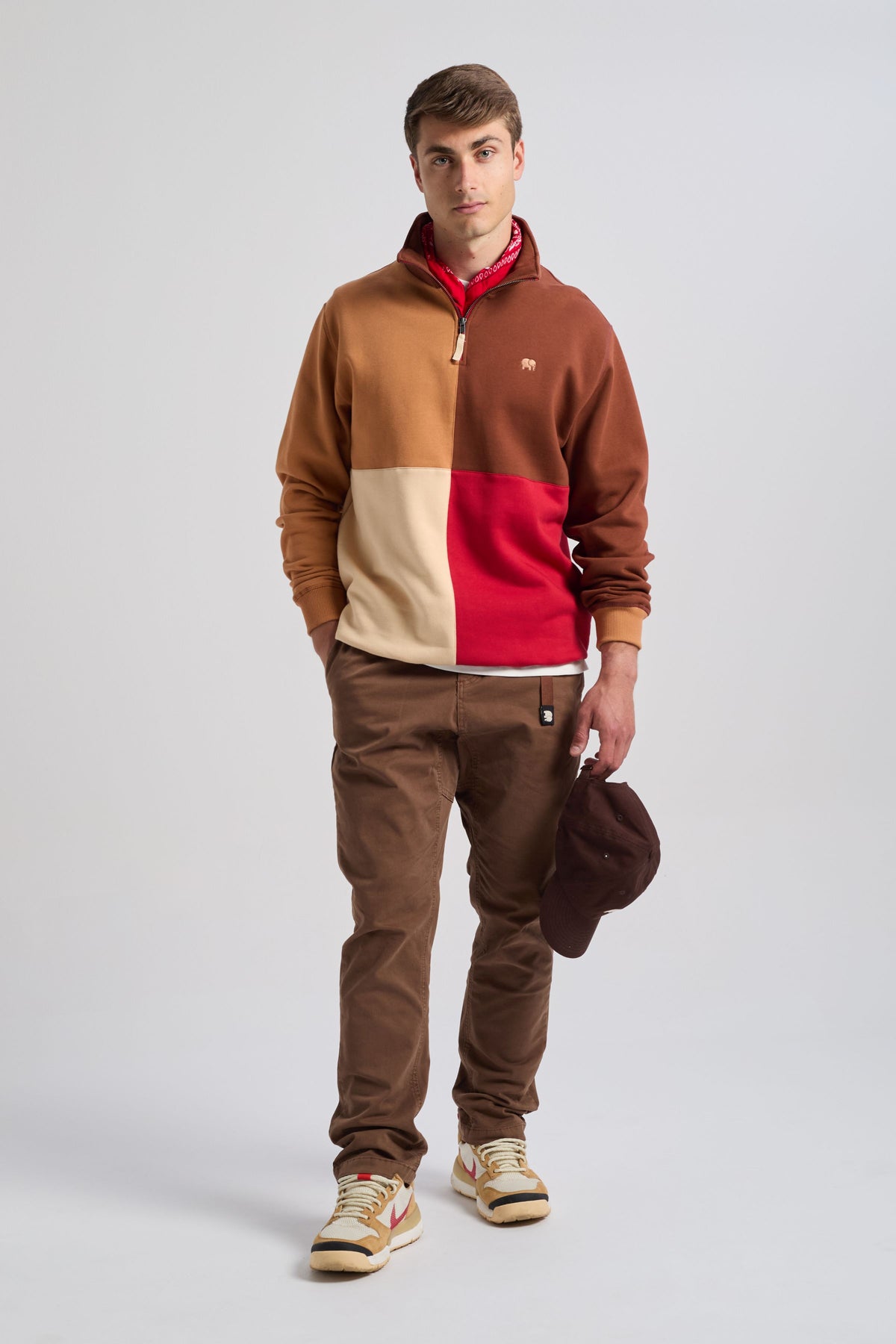 Persona che indossa una Color Block Quarter Zip Sweatshirt in Equestrian Red, con una miscela stilosa di colori tra cui rosso, marrone e beige, abbinata a pantaloni marroni e con un cappellino marrone in mano | TP_IMAGE_TYPE=MODEL_FRONT