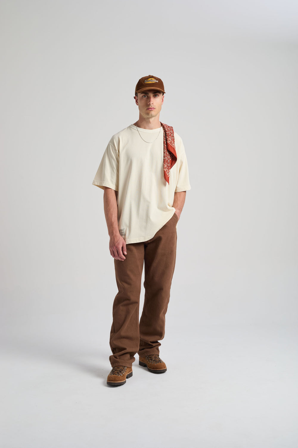 Personne portant un t-shirt oversize blanc antique, stylisé avec un bandana, associé à un pantalon marron et des bottes, debout sur un fond neutre. | TP_IMAGE_TYPE=MODEL_FRONT