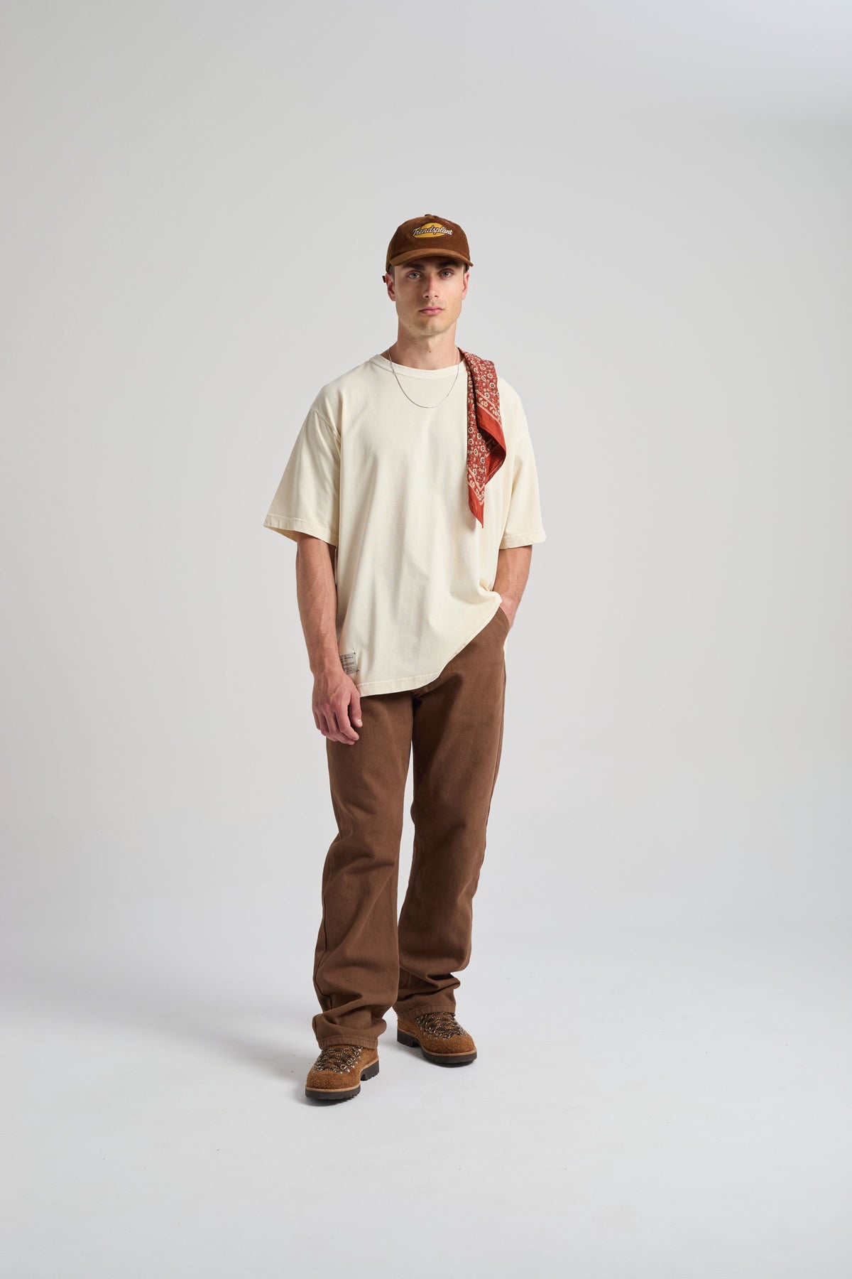 Personne portant un t-shirt oversize blanc antique, stylisé avec un bandana, associé à un pantalon marron et des bottes, debout sur un fond neutre. | TP_IMAGE_TYPE=MODEL_FRONT