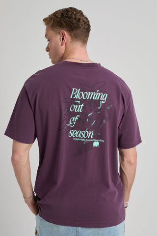 Person wearing a Bloom T-Shirt in Winter Bloom purple, showing the back with a graphic print that reads "Blooming out of season." | TP_IMAGE_TYPE=MODEL_BACK