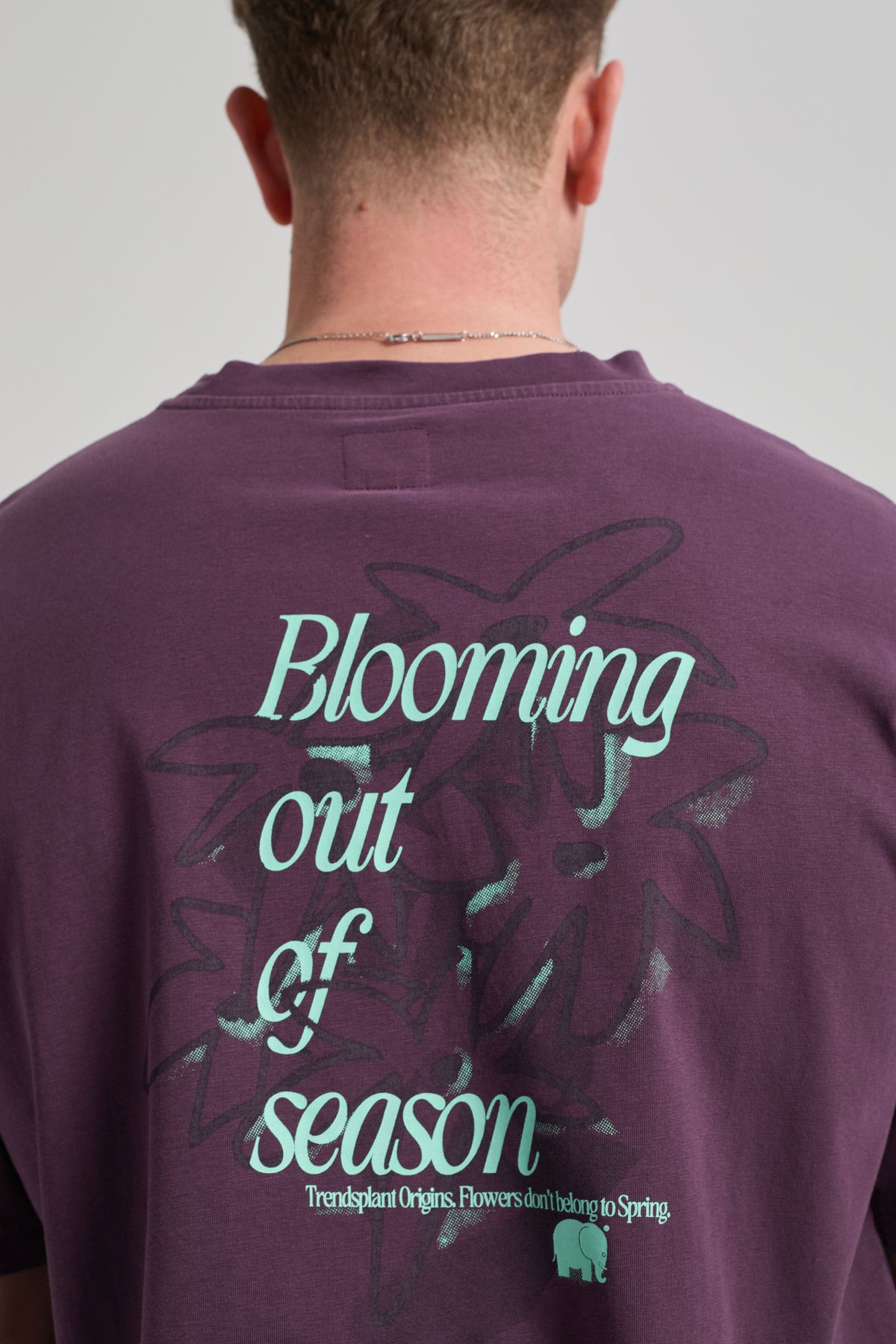 Person wearing the Bloom T-Shirt Winter Bloom, showing the back with a bold graphic design in teal text on a deep purple fabric. | TP_IMAGE_TYPE=MODEL_BACK