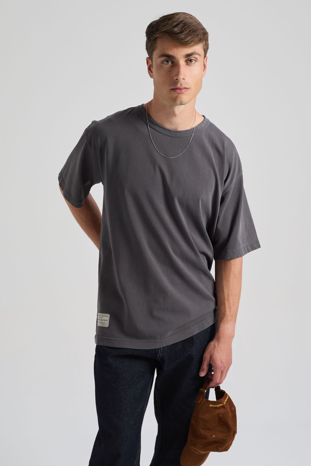 Persona che indossa una T-shirt Supply Oversized in Forged Iron, abbinata a jeans scuri e con un cappellino marrone in mano | TP_IMAGE_TYPE=MODEL_FRONT
