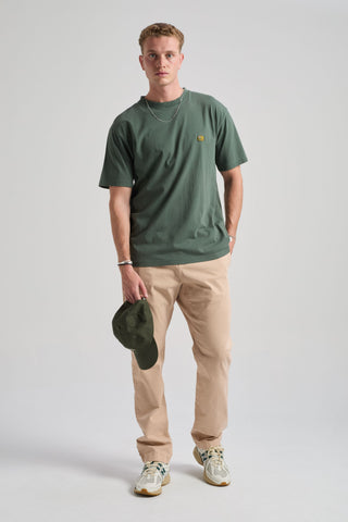 Persona che indossa una t-shirt verde e pantaloni beige, con un cappellino in mano, su sfondo grigio chiaro. TP_IMAGE_TYPE=MODEL_FRONT