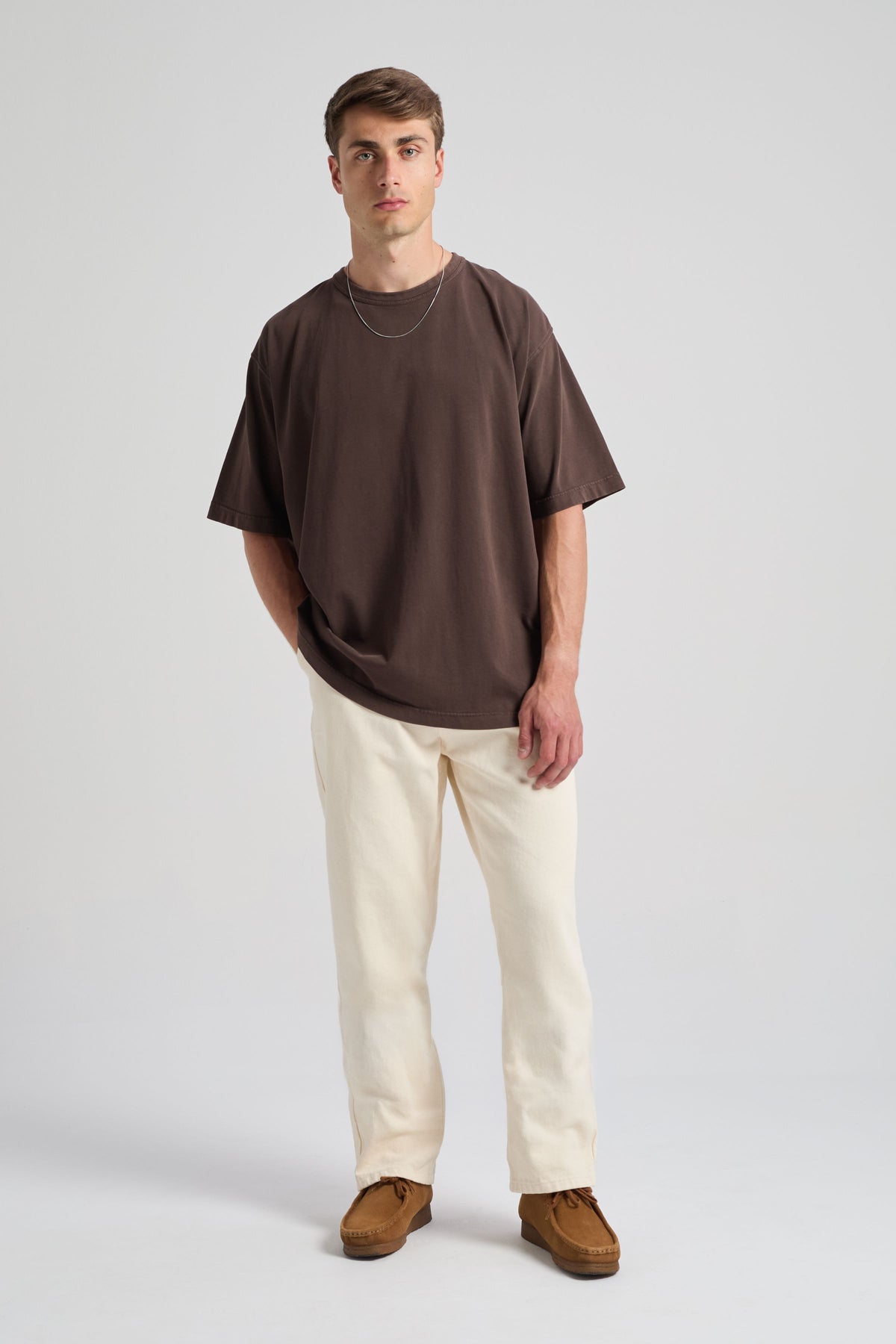 Persona che indossa una t-shirt oversize color chicco di caffè abbinata a pantaloni chiari e scarpe marroni, in piedi davanti a uno sfondo neutro | TP_IMAGE_TYPE=MODEL_FRONT