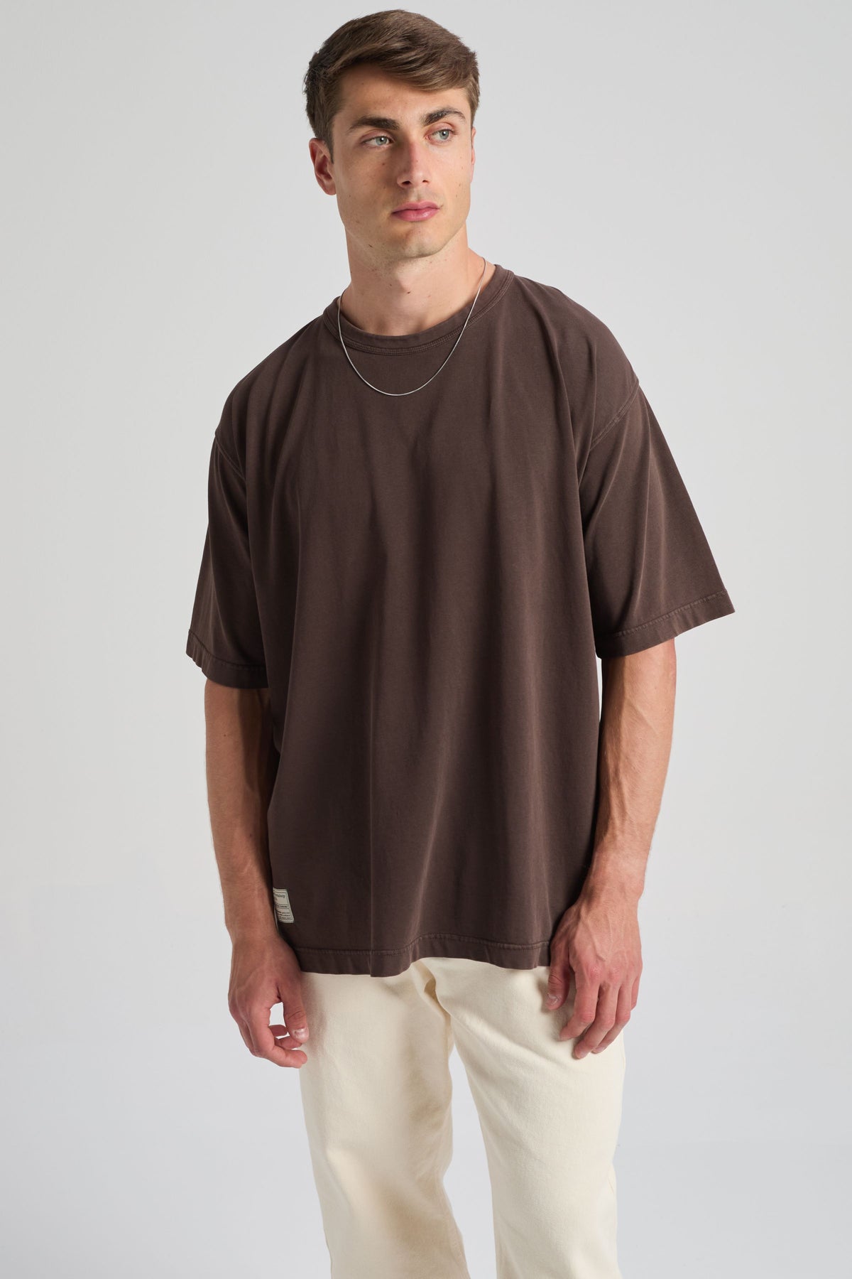Persona che indossa la T-shirt Supply Oversized nel colore Coffee Bean, abbinata a pantaloni chiari, in piedi su uno sfondo neutro | TP_IMAGE_TYPE=MODEL_FRONT
