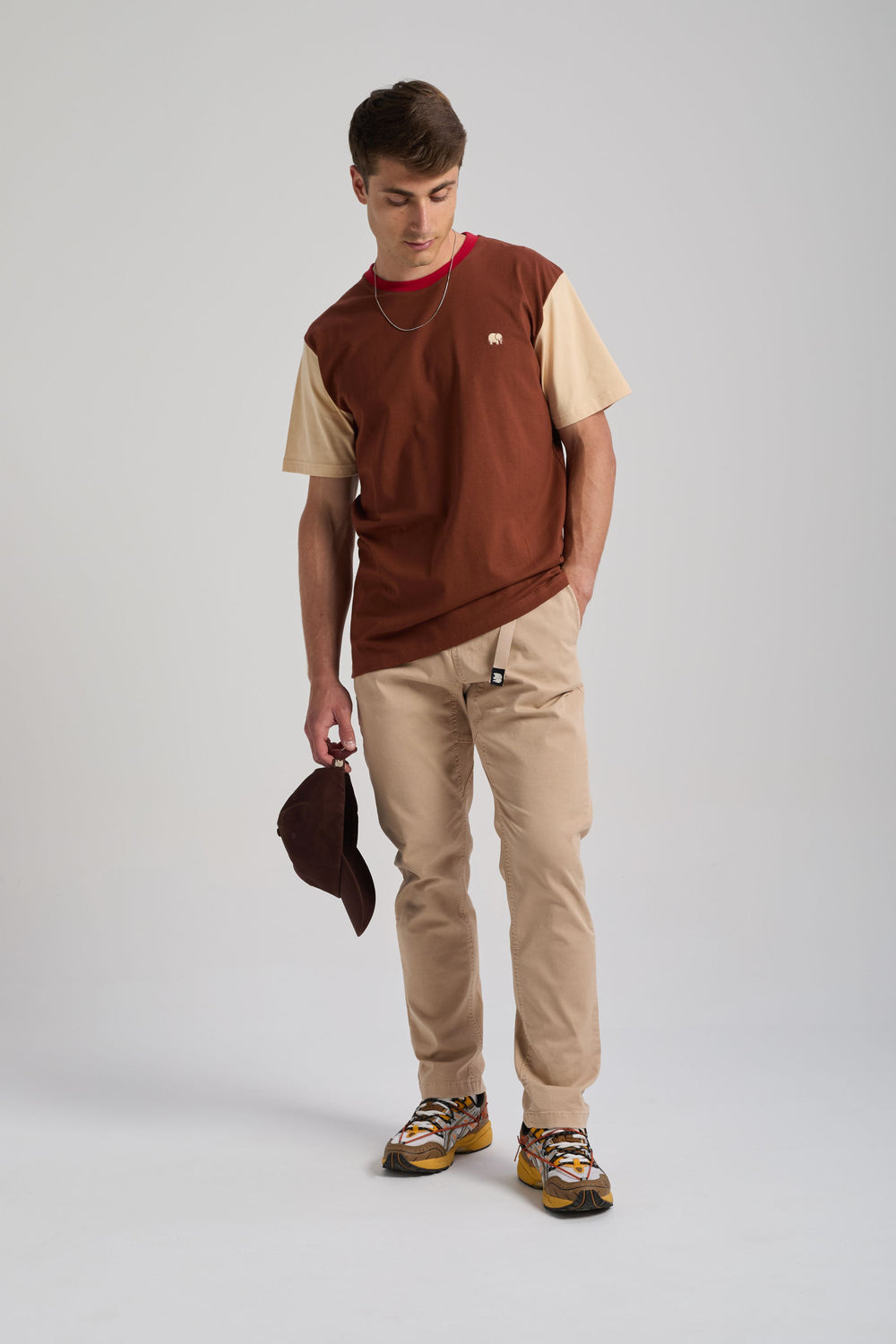 Person wearing a color block t-shirt in cherry mahogany and beige, paired with light-colored pants and holding a brown cap. | TP_IMAGE_TYPE=MODEL_FRONT