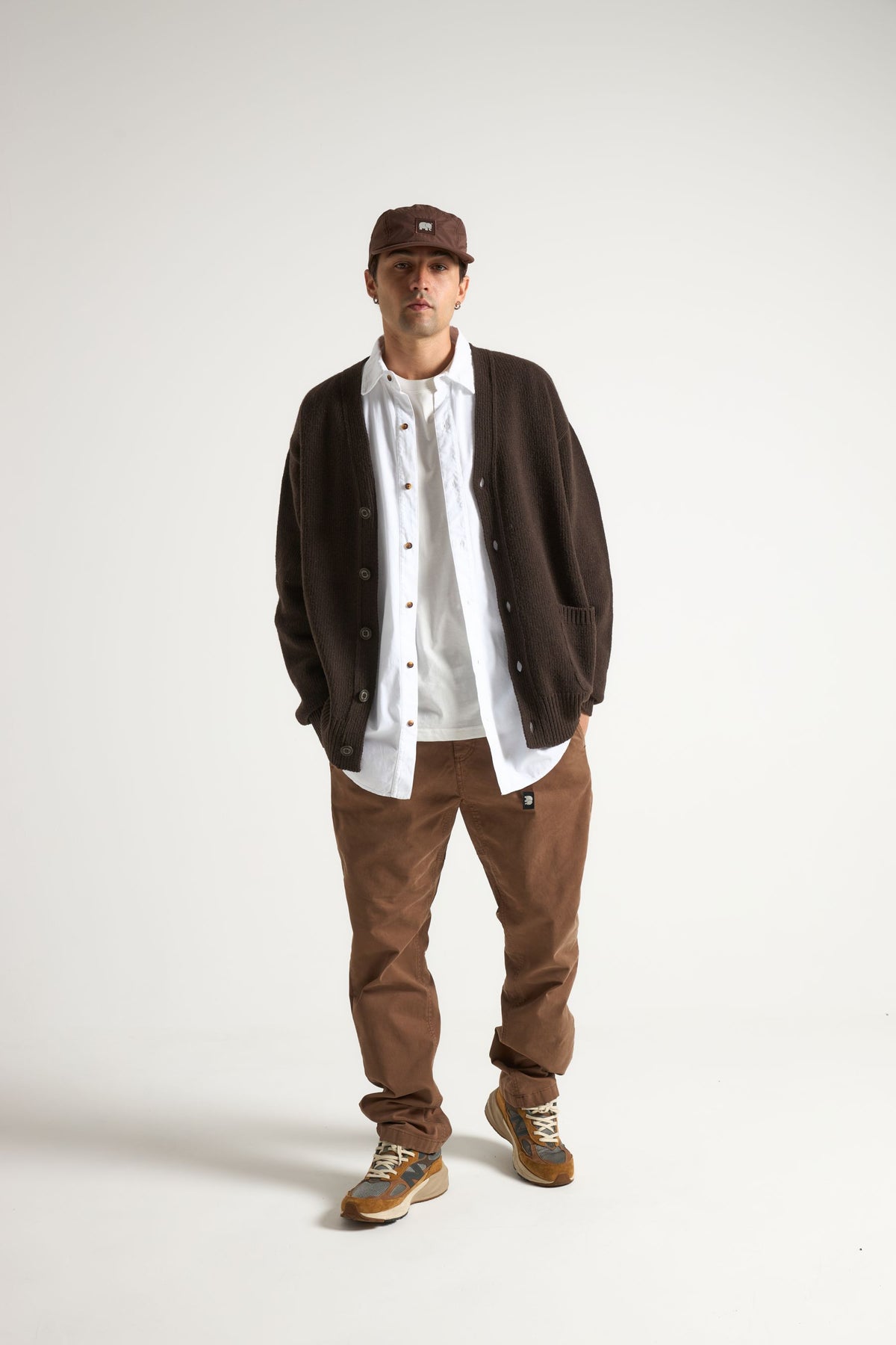 Persona che indossa un cardigan Relleu color chocolate abbinato a una camicia bianca e pantaloni marroni, con cappellino e sneakers, in piedi davanti a uno sfondo neutro | TP_IMAGE_TYPE=MODEL_FRONT
