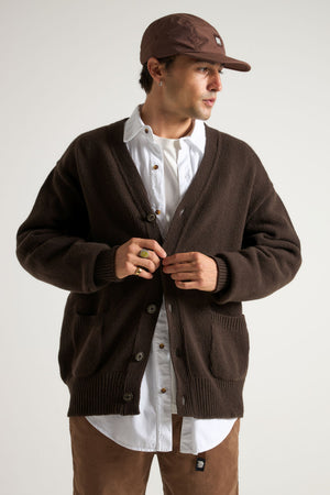Persona che indossa un cardigan Relleu color cioccolato sopra una camicia bianca, in piedi e mentre abbottona il cardigan, con sfondo neutro. | TP_IMAGE_TYPE=MODEL_FRONT