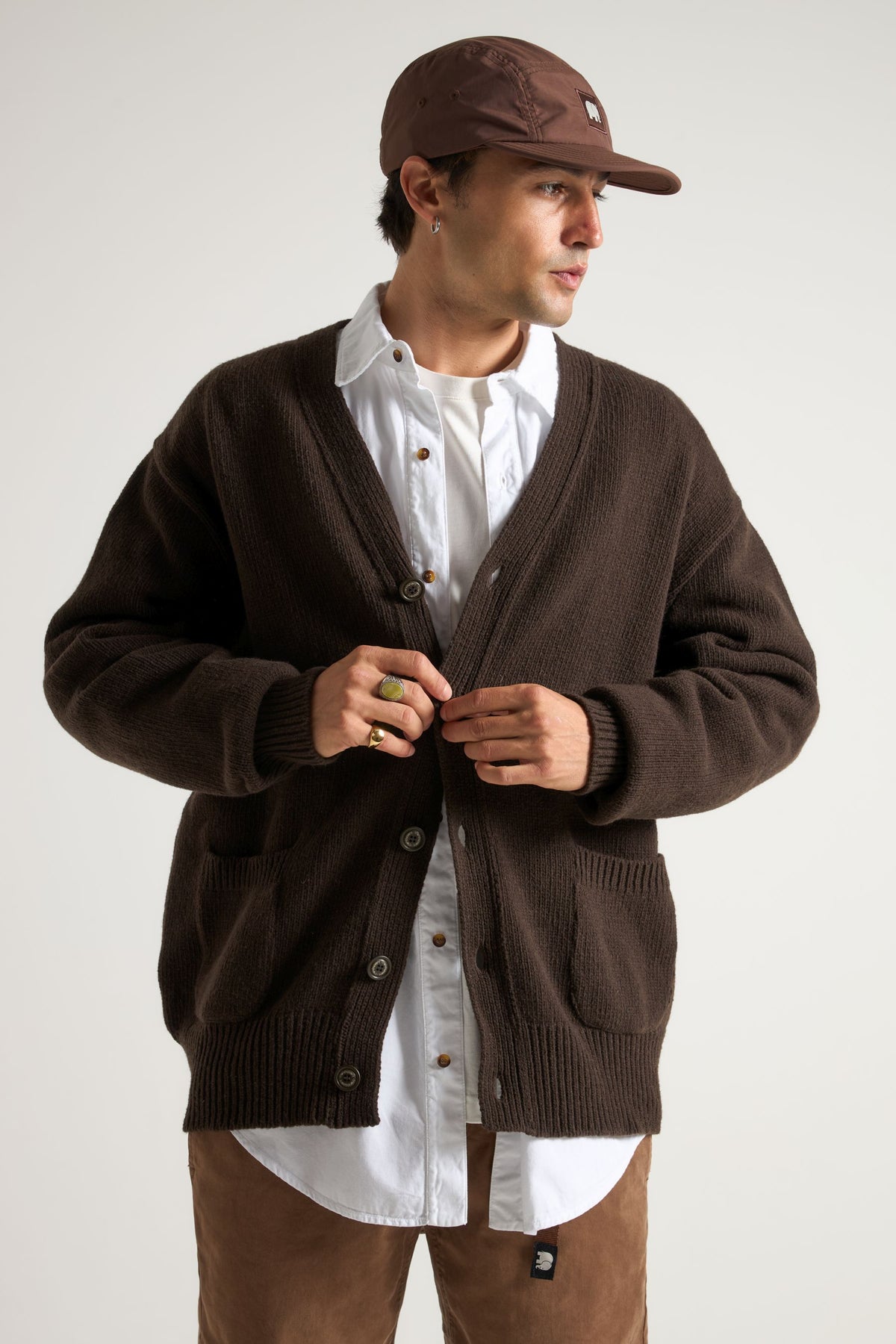 Persona che indossa un cardigan Relleu color cioccolato sopra una camicia bianca, in piedi e mentre abbottona il cardigan, con sfondo neutro. | TP_IMAGE_TYPE=MODEL_FRONT
